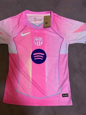 FC Barcelona rosa matchtröja Nike - Snygg FC Barcelona matchtröja i rosa och vitt med lila detaljer. Tröjan har klubbmärke, Nike-logga och Spotify-logga på bröstet. Tillverkad i lätt och ventilerande material, perfekt för fotboll eller träning.