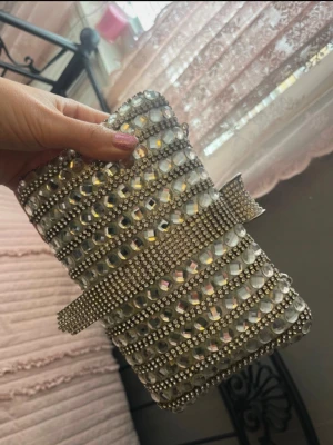 Silver kuvertväska med strassdetaljer - Glittrig kuvertväska i silver med massor av strass och pärlband, samt en dekorativ rem framtill. Väskan har en kedja så den kan bäras på axeln eller i handen. Perfekt accessoar för att sticka ut och ge extra bling till din outfit.