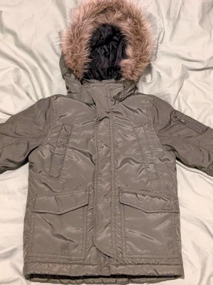 Olivgrön parkas från H&M med fuskpäls - Snygg olivgrön parkas från H&M med varm svart vadderad insida och huva med fuskpäls. Jackan har flera fickor, dragkedja och vindslå framtill samt en ficka på ärmen. Stl. 92, stor i stl.
