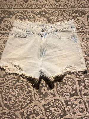 Ljusblå denimshorts med fransar - Säljer ett par ljusblå denimshorts från Defacto med hög midja och råa fransade kanter. Klassisk femficksmodell med knapp och dragkedja framtill. Perfekta för sommaren och riktigt snygga till sneakers eller sandaler.