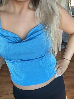 Blå glansig topp från NA-KD - Säljer en snygg blå topp från NA-KD i glansigt material. Toppen har en draperad urringning och är ärmlös med en croppad passform. Perfekt för dig som vill sticka ut med en färgstark och trendig look.