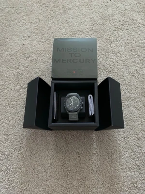 Omega x Swatch Mission to Mercury klocka - Snygg svart Omega x Swatch Speedmaster MoonSwatch Mission to Mercury. Klockan har en mörkgrå boett, svart urtavla med vita detaljer och tachymeter, samt ett grått textilband med Omega-logga. Perfekt för dig som gillar stilrena accessoarer.