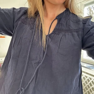 Mörkblå boho-blus med knytband - Snygg mörkblå blus i luftig bomull med avslappnad passform och dekorativa knytband vid halsen. Blusen har subtil spetsdetalj upptill och korta ärmar. Perfekt till jeans eller kjol för en chill look. Passar M!