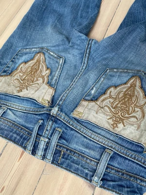 Lågmidjade blåa jeans med broderade bakfickor - Jag säljer ett par utsvängda vintage jeans med låg midja. På bilderna 5-6 kan man se de defekter som finns. Midja:35cm Innerbenslängd:68cm 