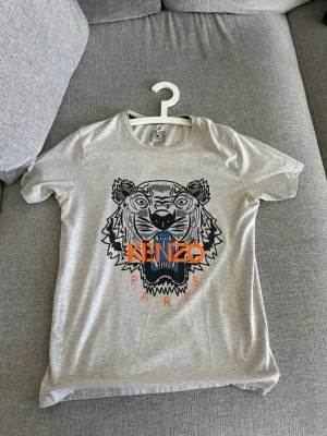 Grå Kenzo t-shirt med tigertryck - Snygg grå t-shirt från Kenzo med ikoniskt tigertryck i svart och blått samt orange text på bröstet. Klassisk rund hals och korta ärmar. Perfekt för dig som vill sticka ut med ett trendigt motiv och en avslappnad passform.