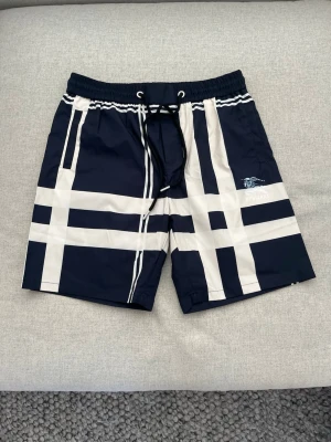 Marinblå badbyxor från Burberry - Snygga marinblå badbyxor från Burberry med klassiskt rutmönster i beige och vitt. De har resårmidja med svart dragsko och sidofickor. Materialet är snabbtorkande och perfekt för stranden eller poolen. Burberry-logga tryckt på ena benet. Passer mer L än XL