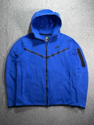 Nike Tech Fleece Full-Zip Hoodie - Märke: Nike Modell: Tech Fleece Windrunner Full-Zip Hoodie Storlek: M Färg: Game Royal (blå) med svarta detaljer Skick: Använd men fint skick – inga fläckar, hål eller skador Material: 53% bomull, 47% polyester Axelbredd: 45 cm Längd: 65 cm