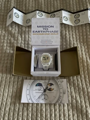 Omega x Swatch Mission to Earthphase Moonshine Gold - Säljer en Omega x Swatch MoonSwatch Mission to Earthphase Moonshine Gold Snoopy. Klockan är vit med biokeramisk boett, vit textilrem och detaljer i blått och guld. Urtavlan har månfaskomplication och tydliga index. Kommer med originalbox och manual.