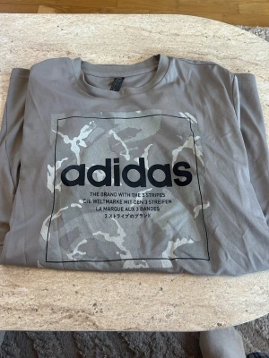 Grå camo t-shirt från Adidas - Snygg grå t-shirt från Adidas med stort logotryck på bröstet och camouflagemönster i vitt och grått. Klassisk rund hals och korta ärmar. Perfekt för dig som gillar streetwear och sportig stil.