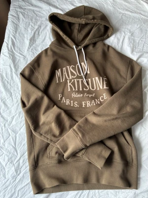 Brun hoodie Maison Kitsuné Paris - Snygg brun hoodie från Maison Kitsuné med vit text på bröstet där det står 'Maison Kitsuné Palais Royal Paris, France'. Hoodien har huva med vita snören och en stor magficka. Tillverkad i mjukt bomullsmaterial, perfekt för en avslappnad streetwear-look.