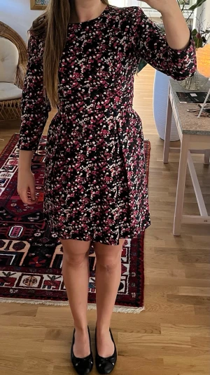 Blommig kortklänning - Kort klänning med svart bas och blommigt mönster i rosa, vitt och rött. Klänningen har lång ärm och rund halsringning. Skön passform med markerad midja och lätt utställd kjol. Perfekt för dig som gillar färg och mönster!