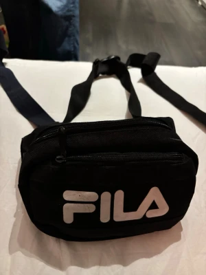 Svart magväska från FILA - Snygg svart magväska från FILA med justerbart band och två dragkedjefack. Väskan har stor vit logga framtill och är tillverkad i slitstarkt tygmaterial. Perfekt för att bära det viktigaste när du är på språng.