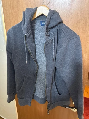 Mörkgrå hoodie från Icon Sports - Mörkgrå hoodie med dragkedja från Icon Sports. Utrustad med stor huva och dragsko, samt två rymliga fickor framtill. Tillverkad i mjukt och tjockt material som känns riktigt skönt mot huden. Perfekt för dig som gillar en enkel och stilren look.