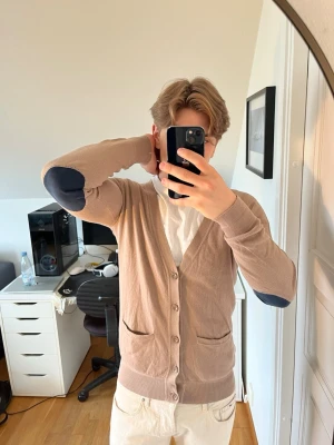 Beige cardigan från Somewhere - Stilren beige cardigan med knappar framtill och två fickor. Tröjan har mörkblå armbågslappar som ger en snygg kontrast. Hör gärna av dig vid frågor!
