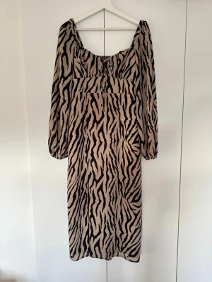 Zebramönstrad klänning - Snygg klänning med zebramönster i beige och svart. Klänningen har långa, lösa ärmar och en snygg snörningsdetalj vid urringningen. Storlek S. 