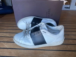 Valentino Garavani vita sneakers - Säljer ett par vita sneakers från Valentino Garavani med en bred svart remsa över sidan. Skorna har klassisk låg profil, vita snören och detaljer i läder. Perfekta för dig som gillar stilrena och exklusiva sneakers.