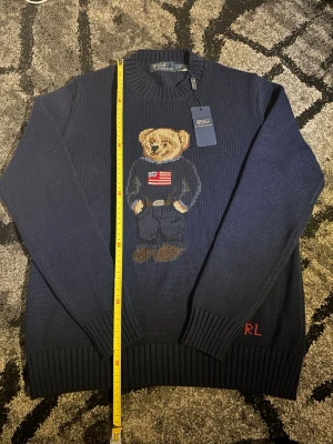 Mörkblå stickad tröja Polo Bear - Mörkblå stickad tröja från Polo Ralph Lauren med den ikoniska Polo Bear på bröstet, klädd i jeans och tröja med amerikanska flaggan. Tröjan har ribbade muddar och RL-broderi vid nederkanten. Perfekt för dig som gillar klassisk preppy stil. Står M fast passar S