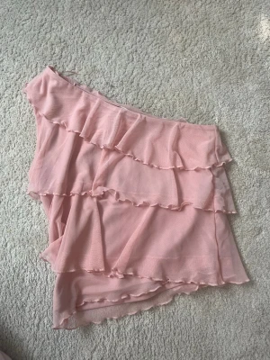 Rosa volangkjol i chiffong - Supersöt rosa off shoulder tröja med flera lager volanger och asymmetrisk form. Tröjan är gjord i ett tunt, lätt material som ger ett flowy intryck. Perfekt nu till sommaren