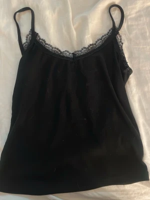Svart linnetopp med spetskant - Snygg svart linnetopp med tunna axelband och dekorativ spetskant längs urringningen. Perfekt att styla lager på lager eller bära som den är. Enkel och stilren design som passar till mycket.