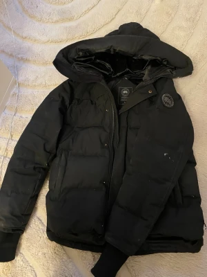 Svart dunjacka från Canada Goose - Säljer en stilren Canada Goose i svart. Använd men fortfarande i okej skick, håller värmen riktigt bra och är perfekt för vintern. Jackan har visst slitage med små hål samt dragskon i huvan som har släppt lite, vilket syns på bilderna och är inräknat i priset.