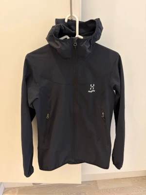 Svart vindjacka från Haglöfs - Snygg svart vindjacka från Haglöfs med hel dragkedja framtill, två fickor med dragkedja och justerbar huva. Jackan har minimalistisk design och diskret logga på bröstet. 