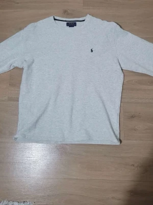 Ljusgrå tröja från Polo Ralph Lauren - Ljusgrå långärmad tröja från Polo Ralph Lauren med klassisk rund halsringning och den ikoniska broderade loggan på bröstet. Tröjan har en enkel och stilren design, perfekt för en avslappnad look. Materialet känns mjukt och bekvämt.