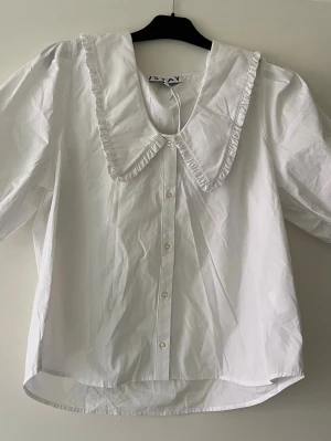 Vit blus med stor krage från STAY - Söt vit blus från STAY med stor dekorativ krage och volangkant. Blusen har knäppning framtill och är tillverkad i ett lätt bomullsliknande material. Perfekt för dig som gillar trendiga detaljer och en romantisk look. I storlek XL