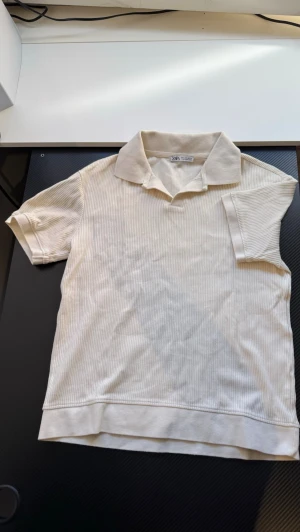 Beige ribbad pikétopp från Zara - Säljer en beige ribbad pikétopp från Zara med korta ärmar och klassisk krage. Toppen har diskreta ränder i materialet och är tillverkad i mjuk bomull, perfekt för en clean och chill look.