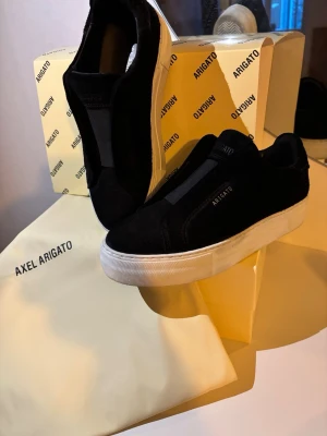 Svarta sneakers från Axel Arigato - Stilrena svarta sneakers från Axel Arigato med tjock vit sula och minimalistisk design. Ovandelen är i mocka och textil, utan snörning för en clean look. Diskret logga på sidan och plös. Perfekta för dig som gillar enkel och modern stil.