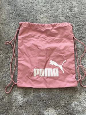 Rosa gympåse från Puma - Säljer en rosa gympåse från Puma med vitt logotryck framtill. Väskan har dragskostängning och tunna snören som axelband. Perfekt för träning eller att bära med sig det viktigaste. Tillverkad i lätt tygmaterial. Ett litet hål men inte så det påverkar innehållet.