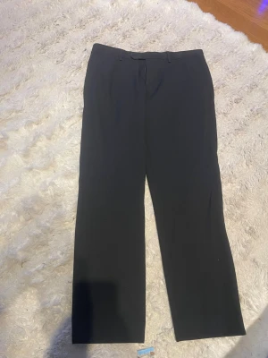 Svarta raka kostymbyxor 34-30 - Säljer ett par klassiska svarta kostymbyxor med rak passform och bälteshällor. Byxorna har diskreta sidofickor och stängs med dragkedja och knapp. Perfekta för en stilren och clean look. Materialet känns mjukt och följsamt. 34-30