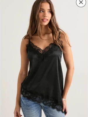 Svart linnetopp med spets - Snygg svart linnetopp i satinliknande material med tunna axelband och spetsdetaljer längs ringning och nederkant. Perfekt att styla med jeans eller kjol🤩Det är helt nytt och oanvänt med prislappen kvar! Ord. pris 399 kr
