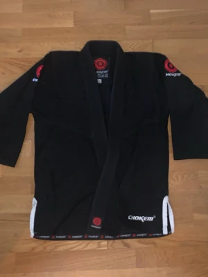 Svart BJJ gi jacka, bälte och byxor från Chokem - Säljer en svart BJJ gi jacka från Chokem med röda och vita detaljer. Jackan har broderade loggor på axlarna och vid kragen samt förstärkta sömmar. Perfekt för brasiliansk jiu-jitsu och grappling.