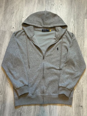 Grå zip hoodie från Polo Ralph Lauren - Snygg grå hoodie med dragkedja från Polo Ralph Lauren. Klassisk design med huva och dragsnören, samt den ikoniska loggan broderad på bröstet. Tillverkad i mjukt bomullsmaterial och har ribbade muddar vid ärmar och nederkant. Perfekt för en chill och stilren look.