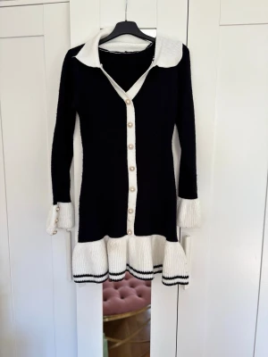 Svart och vit stickad klänning - Snygg svart stickad klänning med vita detaljer, krage och knappar framtill. Klänningen har lång ärm, volangkant nertill och ribbade muddar. Perfekt för dig som gillar en klassisk men trendig look.