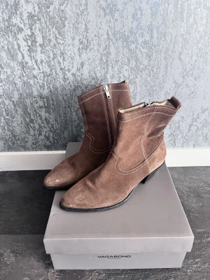 Bruna mockastövletter från Vagabond - Snygga bruna stövletter i mocka från Vagabond med låg klack och spetsig tå. Dragkedja på sidan och dekorativa sömmar ger en cool westernkänsla. Perfekta för dig som vill ha en stilren och trendig look.