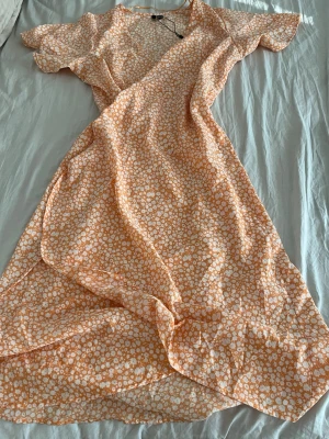 Blommig omlottklänning i orange och vit - Säljer en lång omlottklänning med blommigt mönster i orange och vitt. Klänningen har korta ärmar, v-ringning och volangdetaljer längs kanterna. Perfekt för sommarens alla tillfällen!