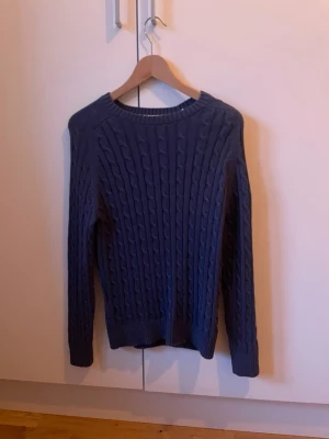 Superdry Mörkblå kabelstickad tröja - PRIS KAN DISKUTERAS. Säljer en mörkblå kabelstickad tröja med rund hals och långa ärmar. Tröjan har ett klassiskt flätmönster och ribbade muddar vid ärmslut och nederkant. Perfekt för dig som gillar stilrena och tidlösa plagg.