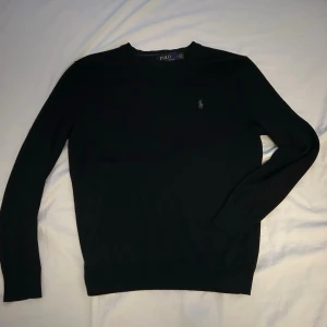 Svart stickad tröja Polo Ralph Lauren - Stilren svart stickad tröja från Polo Ralph Lauren i slim fit-modell. Tillverkad av tvättbar merinoull och har rund halsringning med en grå logga på bröstet. Inga defekter.