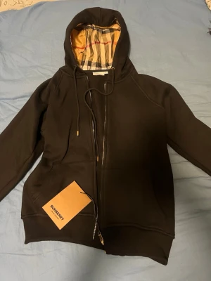 Svart hoodie från Burberry - Snygg svart hoodie från Burberry med dragkedja och klassiskt rutigt foder i huvan. Hoodien har fickor framtill, justerbar dragsko och guldfärgade detaljer på snöret. Perfekt för dig som vill ha en stilren och exklusiv look.