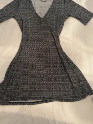 Mönstrad klänning från Gina Tricot - Kort klänning från Gina Tricot i svart och grått med vågigt mönster. Klänningen har v-ringning och trekvartsärm, tillverkad i mjukt och stretchigt material som känns skönt mot huden. Perfekt för dig som gillar en avslappnad men ändå snygg stil.