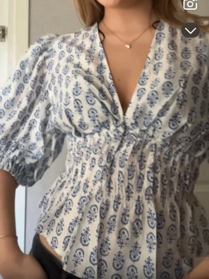 Vit blus med blått mönster - Söt vit blus med blått paisleymönster och djup v-ringning. Blusen har puffiga trekvartsärmar och smockad midja som ger en feminin siluett. Tillverkad i ett lätt och luftigt tyg, perfekt för varma dagar. Passar till både jeans och kjol. Jag har storlek M på denna och är vanligtvis XS/S. Använd 1 gång