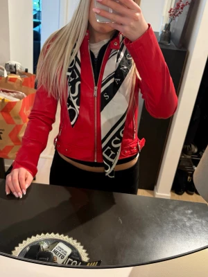 Röd skinnjacka från Guess - Snygg röd skinnjacka från Guess med silverfärgade dragkedjor och detaljer. Jackan har en kort, figurnära modell med axelvaddar och bälte i midjan. Perfekt för dig som vill sticka ut och addera färg till din outfit.
