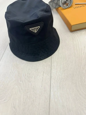 Svart bucket hat från Prada - Snygg svart bucket hat från Prada med det klassiska trekantiga logotypmärket framtill. Hatten är tillverkad i ett glansigt nylonmaterial som ger en modern och trendig look. Perfekt accessoar för att lyfta din outfit och ge en exklusiv touch.