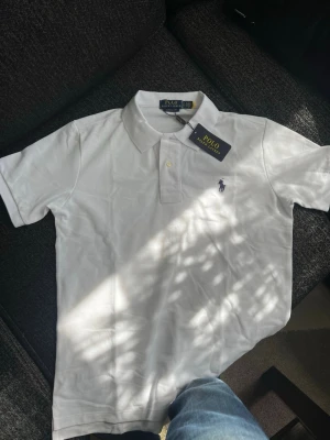 Vit pikétröja från Polo Ralph Lauren - Klassisk vit pikétröja från Polo Ralph Lauren med krage och två knappar framtill. Tröjan har den ikoniska broderade loggan på bröstet och är tillverkad i mjuk bomull. Perfekt för en clean och stilren look.