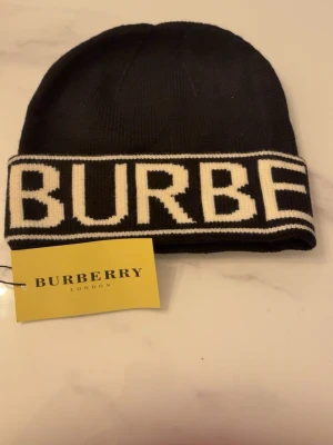 Svart mössa från Burberry - Snygg svart mössa från Burberry med bred vit kant och stor logotyp i vitt runt om. Klassisk ribbstickad design som ger en clean och modern look. Perfekt accessoar för dig som vill ha något stilrent och trendigt. Materialet är mjukt och bekvämt.