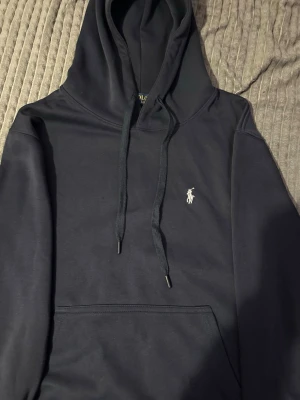 Mörkblå hoodie från Polo Ralph Lauren - Snygg mörkblå hoodie från Polo Ralph Lauren med klassisk huva och dragsnören. Har en broderad vit logga på bröstet och en stor magficka framtill. Perfekt för en chill och stilren look. Materialet känns mjukt och bekvämt.