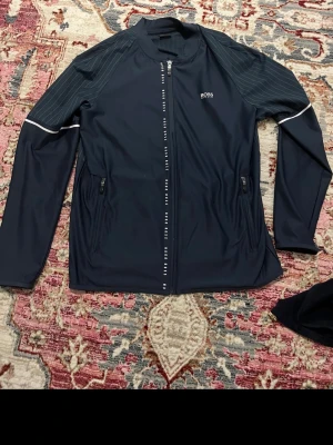 Sporthoodie Hugo Boss - Snygg svart sportjacka från Hugo Boss med dragkedja framtill och diskreta vita detaljer. Jackan har två fickor med dragkedja och tunna ränder på axlarna. Tillverkad i ett lätt och stretchigt material som passar perfekt för träning eller aktiva dagar.