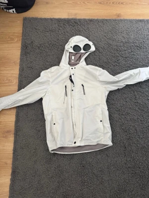 Vit vindjacka med goggles från C.P. Company - Säljer en vit vindjacka från C.P. Company med ikoniska goggles i huvan. Jackan har dragkedja framtill, två bröstfickor med dragkedja och justerbar huva. Perfekt för dig som vill sticka ut med en unik och stilren look.