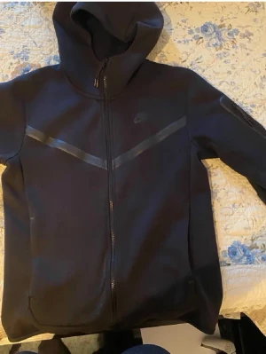 Svart Nike Tech Fleece hoodie - Nike Tech Fleece hoodie i svart med hel dragkedja och huva. Snygga detaljer med svarta band över bröstet och diskret Nike-logga på vänster sida. Tillverkad i mjukt och lätt material som håller värmen utan att bli för tung. Perfekt för en sportig och avslappnad stil.
